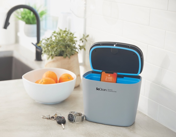Image of Soclean Device on Countertop next to a fruit bowl with oranges in it. Next to the unit infront there is a watch and some keys. The Soclean device is gray, opens at the top and has a blue interior. In this particular pucture there is also a phone inside it. The unit is open, resumably ready for disinfection procedure. The background is a white kitchen backslpash, further back there is a farm style sink under a finely beveled granite countertop the color of beige or sand. In the picture it is proabbly about noon time.